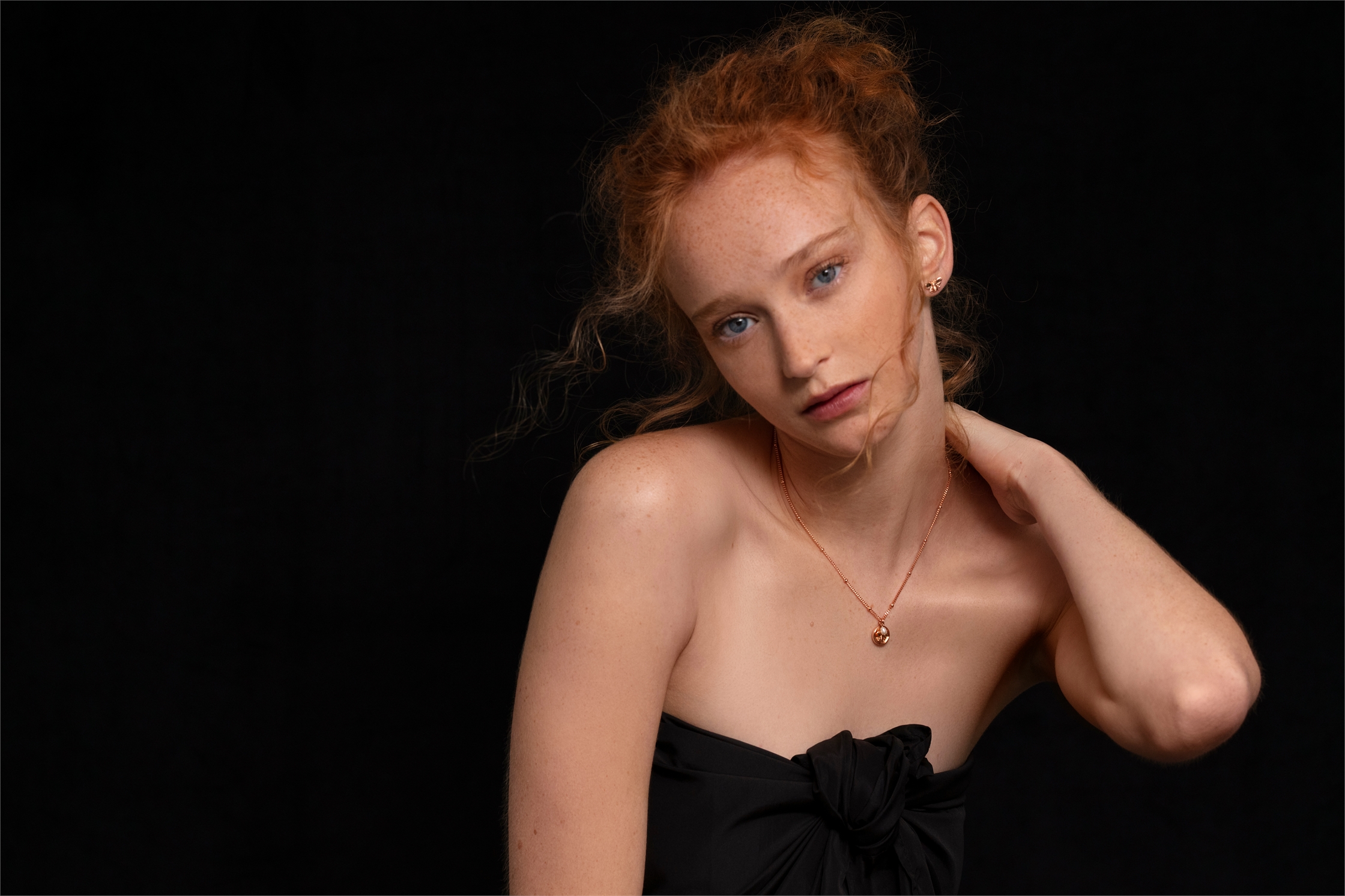 Portrait of a young woman with red hair wearing a black strapless top and gold necklace against a dark background.