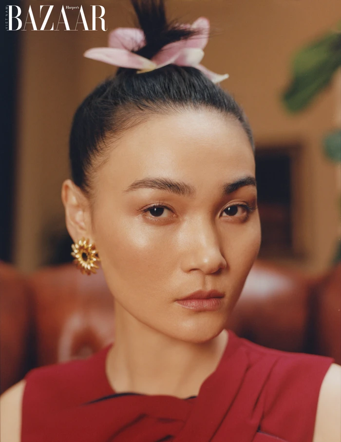 Close-up portrait of an East Asian woman wearing a red top and gold sunburst earrings, with a pink flower tucked into her topknot.