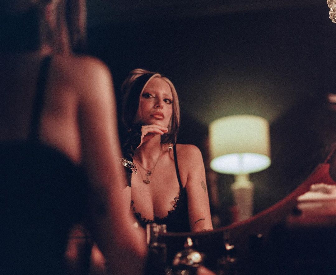 Young woman with blonde streaks poses in black lingerie, reflected in a mirror with a table lamp visible in the background.