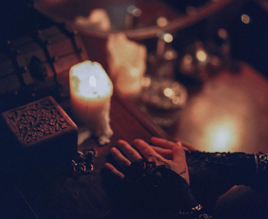 An ornate wooden box sits near a lit candle with a person's hands resting on a table in a dimly lit, indoor setting.