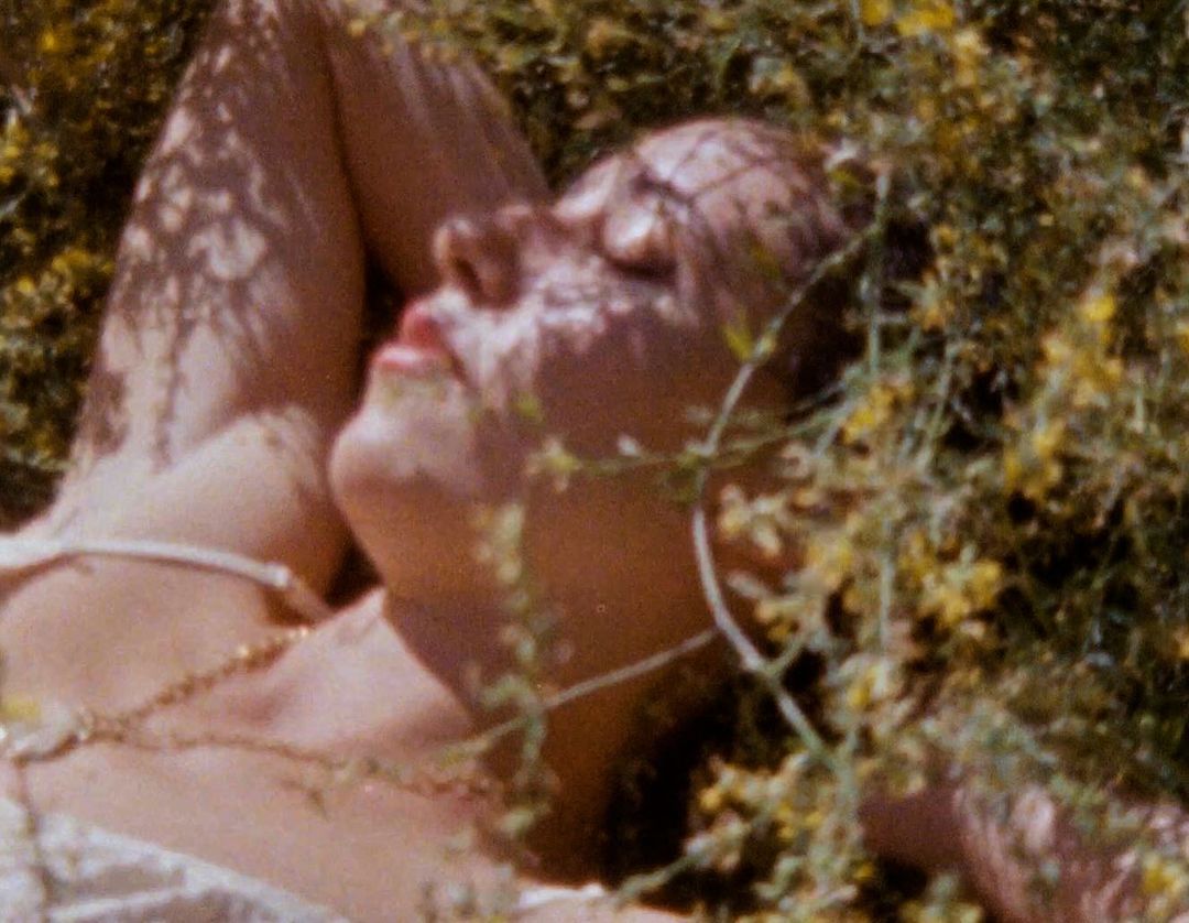 Adult female lies in a field of yellow flowers in dappled sunlight, wearing a gold necklace and a light-colored top with straps.