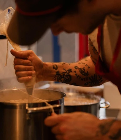 Tattooed cook strains pasta from a large metal pot in a kitchen. Food preparation is evident in this brightly lit indoor scene.
