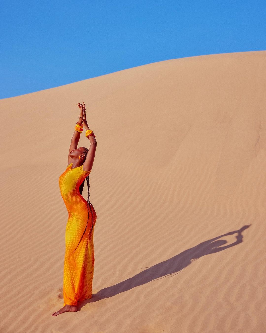 Adult woman in yellow dress with arms raised on sand dune with blue sky in outdoor environment, casting shadow on the ground.