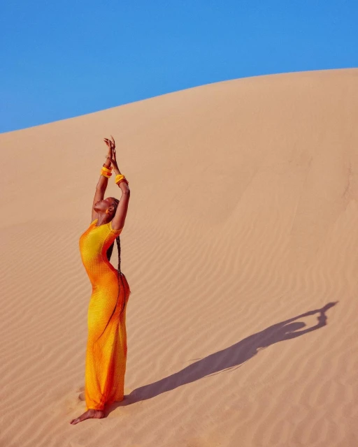 Adult woman in yellow dress with arms raised on sand dune with blue sky in outdoor environment, casting shadow on the ground.