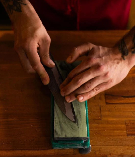 Close-up of hands sharpening a knife on a sharpening stone, set on a wooden surface, showcasing the precision and care in culinary tool maintenance.