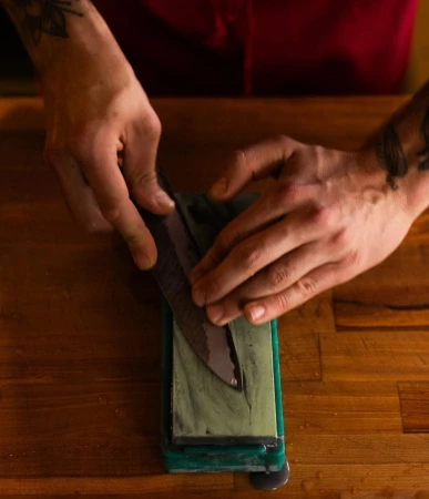 Close-up of hands sharpening a knife on a sharpening stone, set on a wooden surface, showcasing the precision and care in culinary tool maintenance.