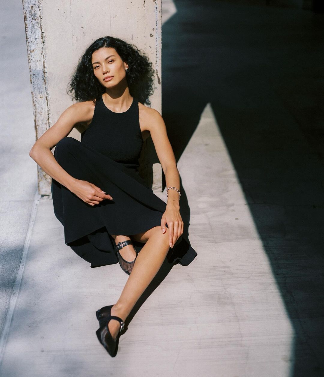 An adult female with curly hair poses seated against a concrete wall, wearing a black dress and Mary Jane shoes in a medium shot.