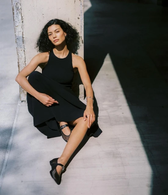 An adult female with curly hair poses seated against a concrete wall, wearing a black dress and Mary Jane shoes in a medium shot.