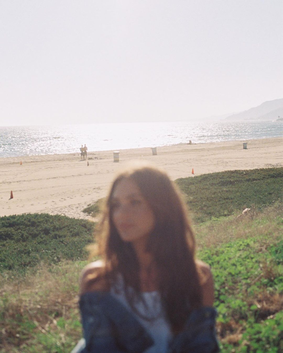Woman with long brown hair stands on a grassy bank overlooking a sandy beach and ocean on a sunny day in an out-of-focus shot.