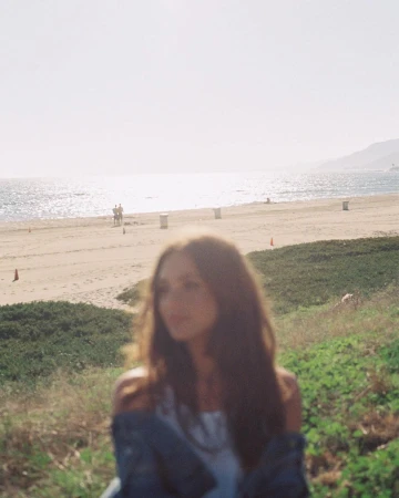 Woman with long brown hair stands on a grassy bank overlooking a sandy beach and ocean on a sunny day in an out-of-focus shot.