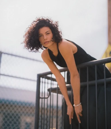 Woman with curly dark hair wearing a black dress leaning against a metal railing outdoors in Los Angeles.