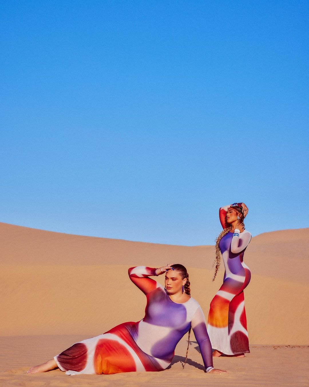 Two women pose in vibrant patterned dresses in a desert landscape under a clear blue sky, creating a striking fashion image.