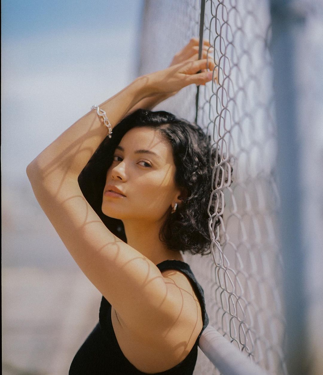 A woman with short curly hair poses in front of a chain-link fence on a sunny day, wearing a sleeveless black top and silver jewelry.