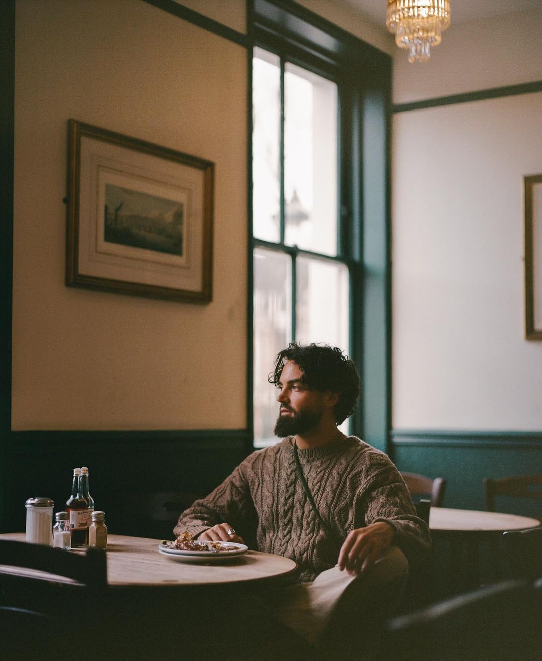 Man in a brown sweater sits at a round table in a restaurant. He is looking off to the side. A chandelier hangs from the ceiling.