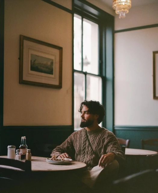Man in a brown sweater sits at a round table in a restaurant. He is looking off to the side. A chandelier hangs from the ceiling.