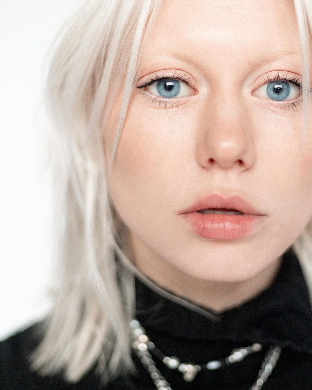 Close-up portrait of a fair-skinned woman with light blonde hair, wearing a black turtleneck and necklace, against a white background.