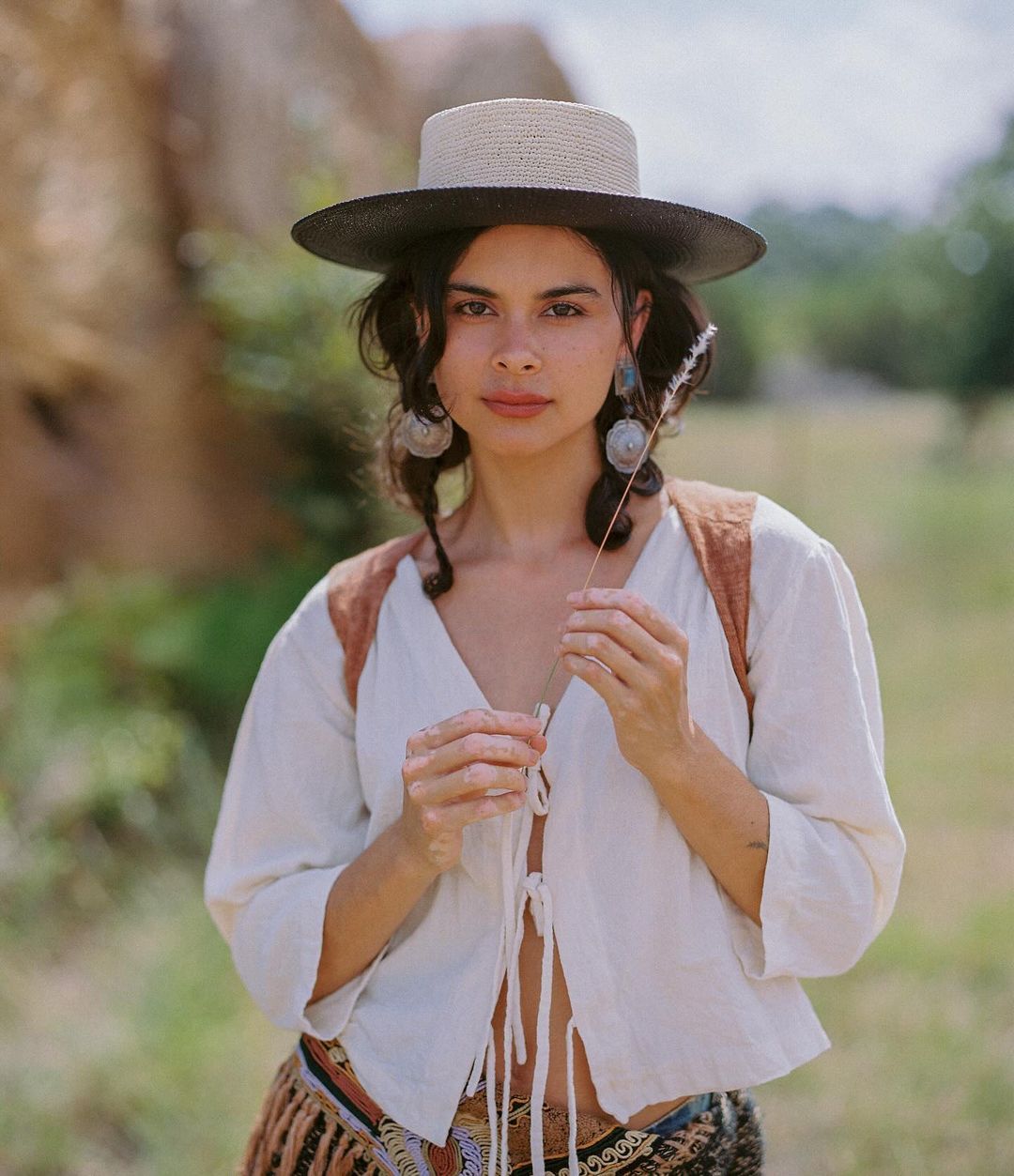 A woman in a brimmed hat stands in a field with a sprig of grass, wearing a white blouse, against a background of rocks and greenery.