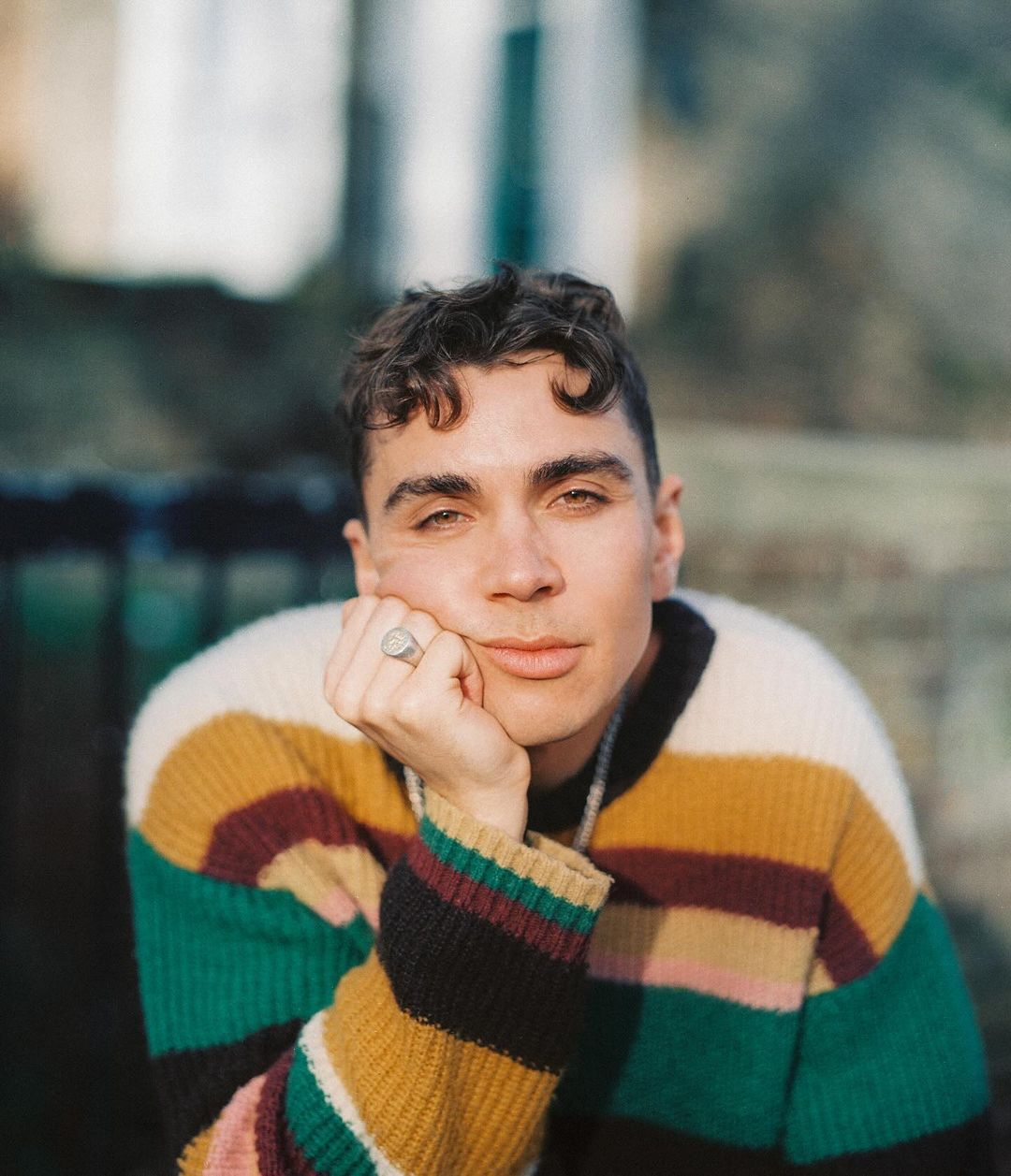 Portrait of a young adult male with curly hair wearing a striped sweater sitting outdoors, looking directly at the camera with a neutral expression.