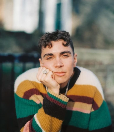 Portrait of a young adult male with curly hair wearing a striped sweater sitting outdoors, looking directly at the camera with a neutral expression.