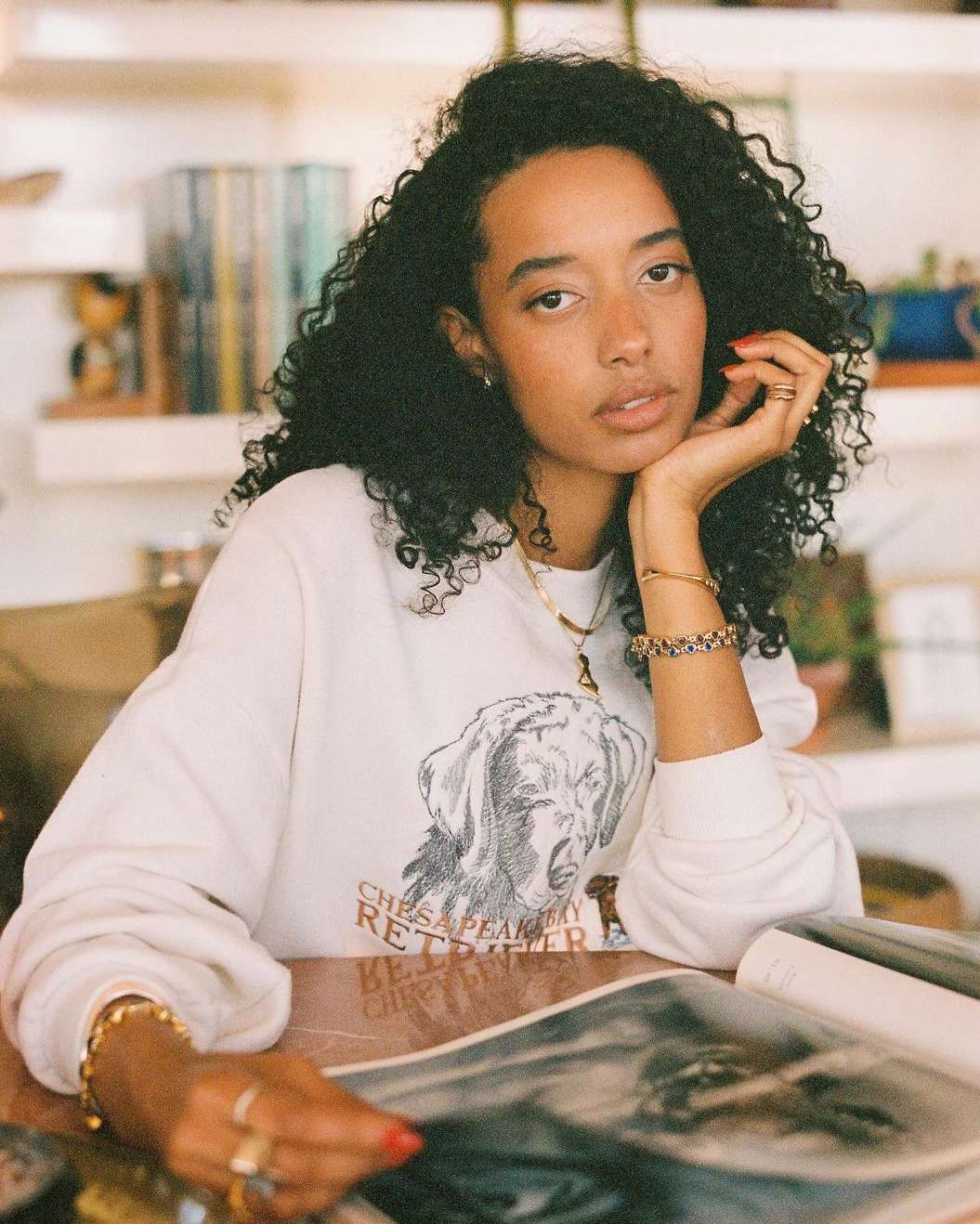 Woman with curly hair wearing a graphic sweatshirt sitting at a table with magazine and jewelry, indoors, in soft, natural light.