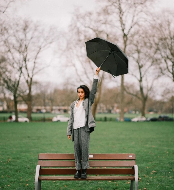 A woman in a grey suit stands on a park bench holding a black umbrella overhead on an overcast day with bare trees in the background.