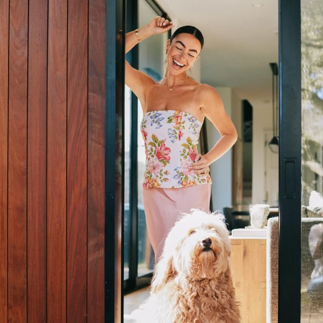 Adult woman in floral top and dog pose near a modern doorway with wood panels; natural light, cheerful expression, and stylish details.