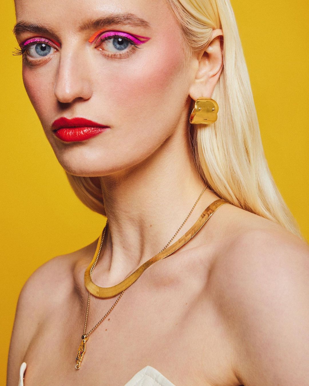 Close-up beauty shot of a woman with blond hair, wearing gold jewelry, bright pink and orange eye makeup, and red lipstick against yellow background.