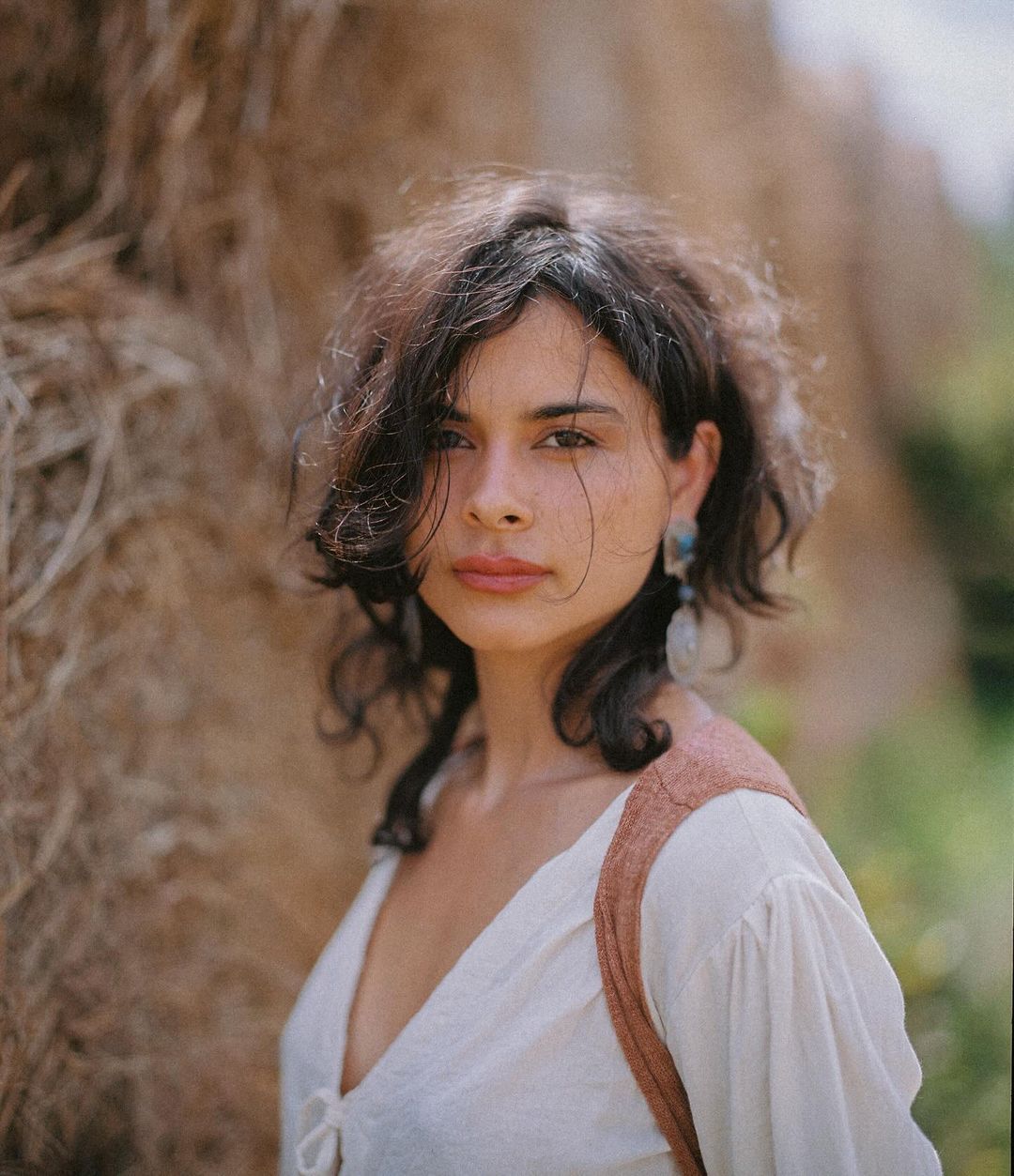 Portrait of a young woman with dark hair in front of a hay bale in an outdoor setting, wearing a white peasant blouse.