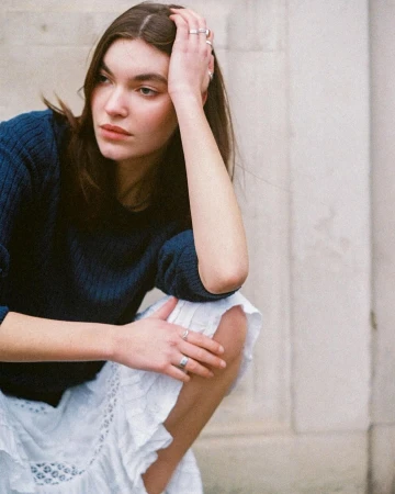 Young woman in a dark blue sweater and white skirt squatting outdoors with hand in hair, photographed by Bethany Reed