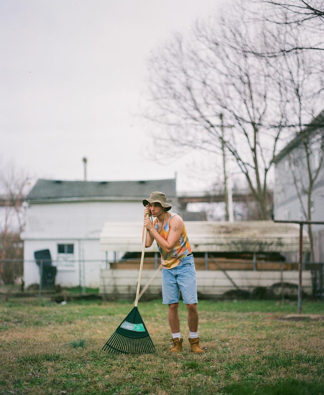 Adult male in yard stands next to rake wearing hat, t-shirt and jean shorts with houses and trees in the background on overcast day.