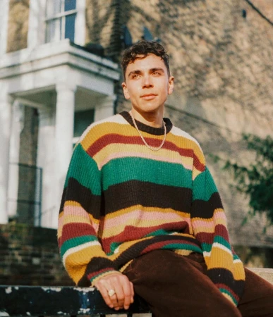 Young man wearing a multi-coloured striped knit sweater posing outdoors near a classic residential building.