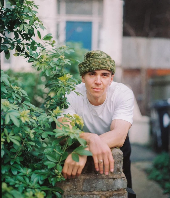 Portrait of a young adult man wearing a beanie and white t-shirt, posing outdoors with greenery, against a white building facade.