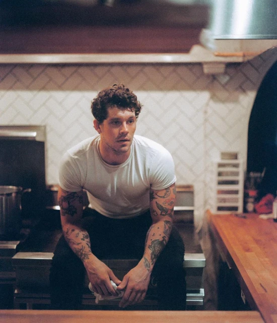Man with tattoos sits in commercial kitchen wearing a white t-shirt and dark pants, looking off-camera. Stainless-steel elements visible.