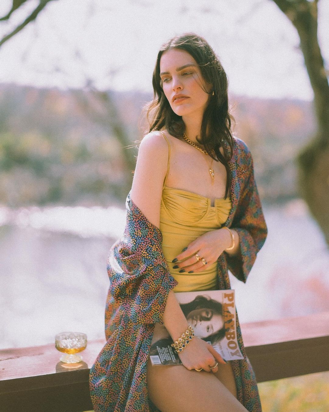 Woman in a gold swimsuit poses with a Playboy magazine on a veranda with a river backdrop; wearing jewelry and a colorful robe.