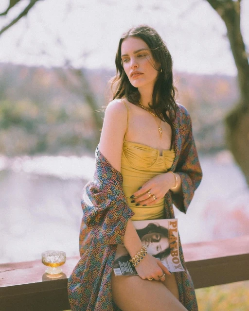 Woman in a gold swimsuit poses with a Playboy magazine on a veranda with a river backdrop; wearing jewelry and a colorful robe.