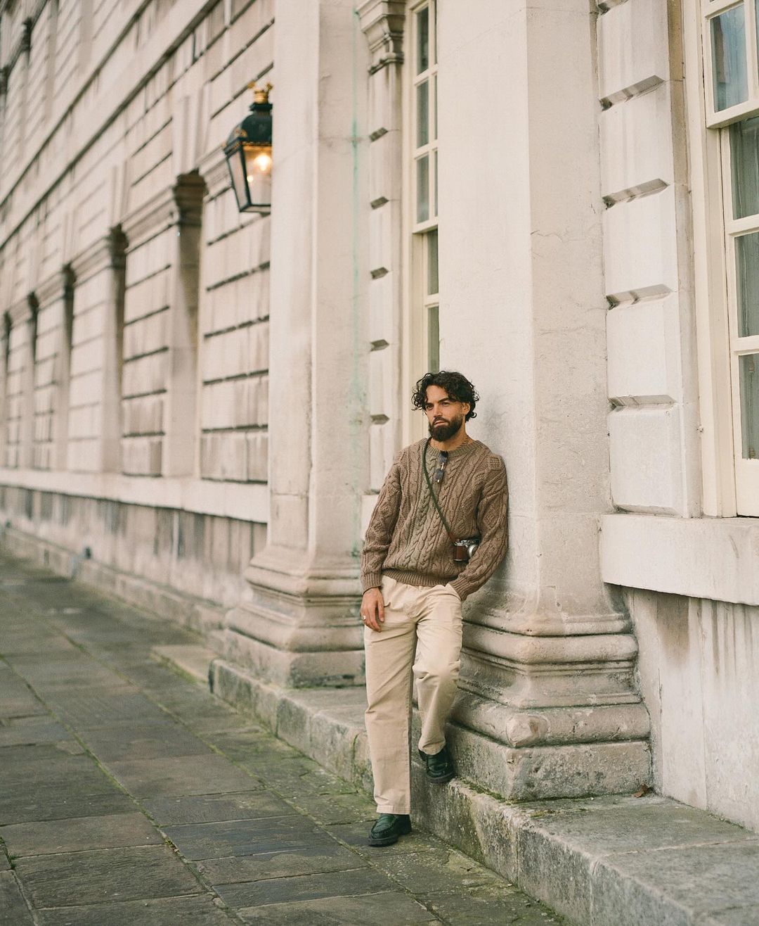 Man stands near building, wearing brown cable-knit sweater, light trousers, and black shoes, with camera on shoulder and relaxed expression.