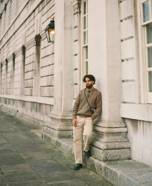 Man stands near building, wearing brown cable-knit sweater, light trousers, and black shoes, with camera on shoulder and relaxed expression.