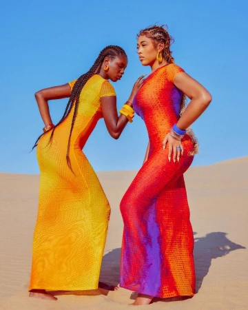 Two models wearing brightly coloured mesh dresses posing on a sandy desert dune under a clear blue sky