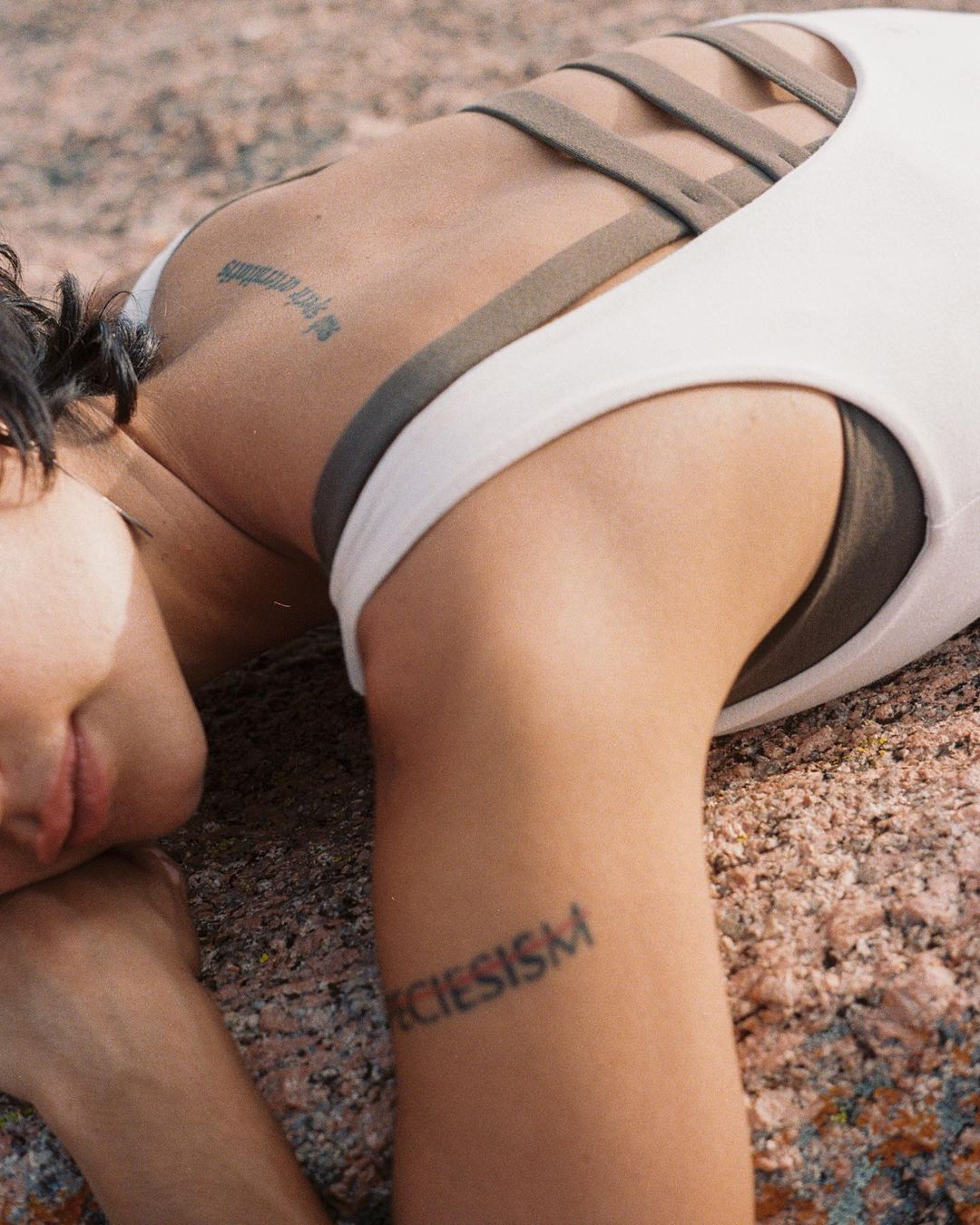 Woman with tattoos lies on rock surface in white top with strap detail, photographed at eye-level in a natural, outdoor setting.