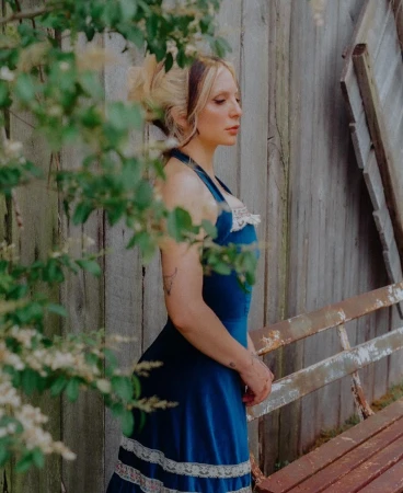 Woman wearing a blue halter dress standing outdoors near a weathered wooden fence and bench, photographed by Bethany Reed.