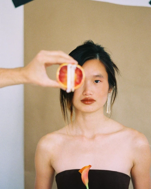 Woman poses with sliced orange and calla lily in a studio setting. The image features a blend of beauty and artistic composition.