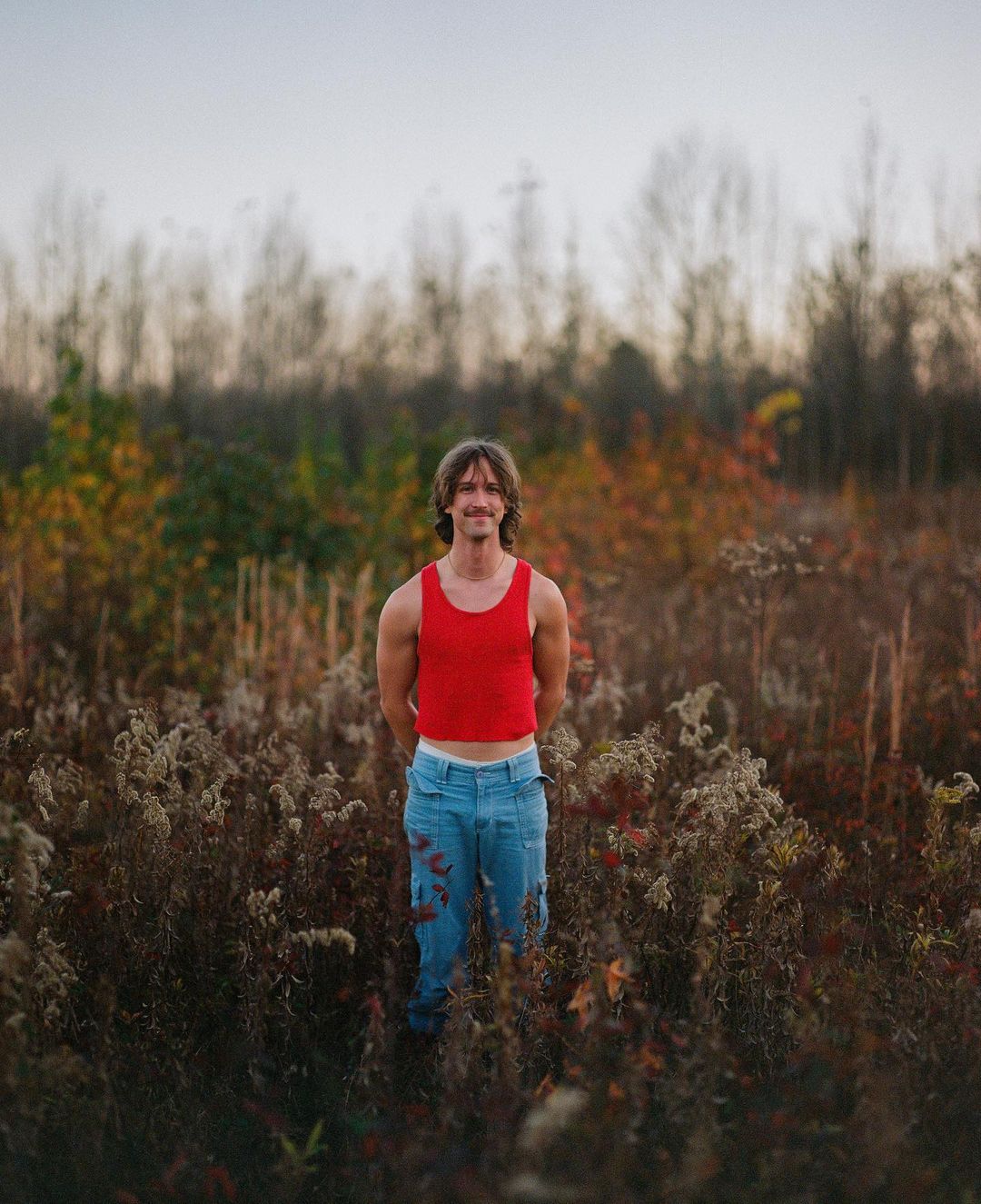 Adult male standing in a field of tall grass and autumn trees wearing a red tank top and blue pants looking at the camera.