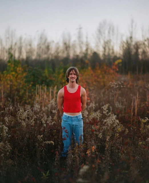 Adult male standing in a field of tall grass and autumn trees wearing a red tank top and blue pants looking at the camera.