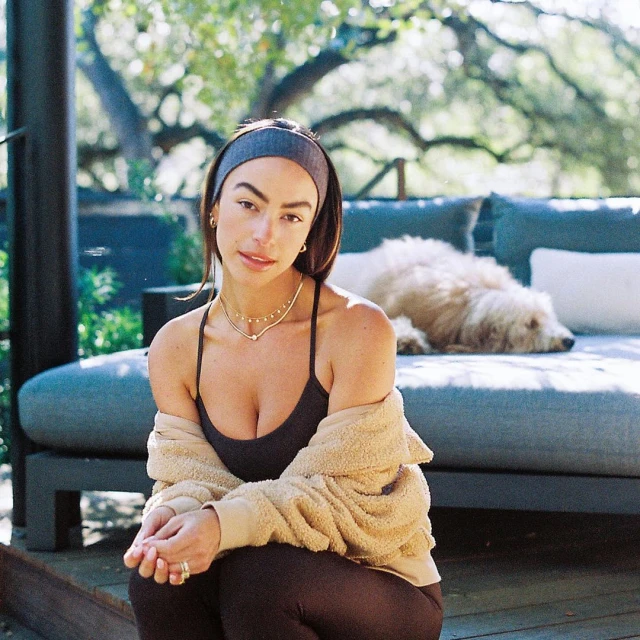 Brunette woman in sportswear sits outside with dog on couch. She wears a headband, tank top, jacket, and leggings in a natural setting.