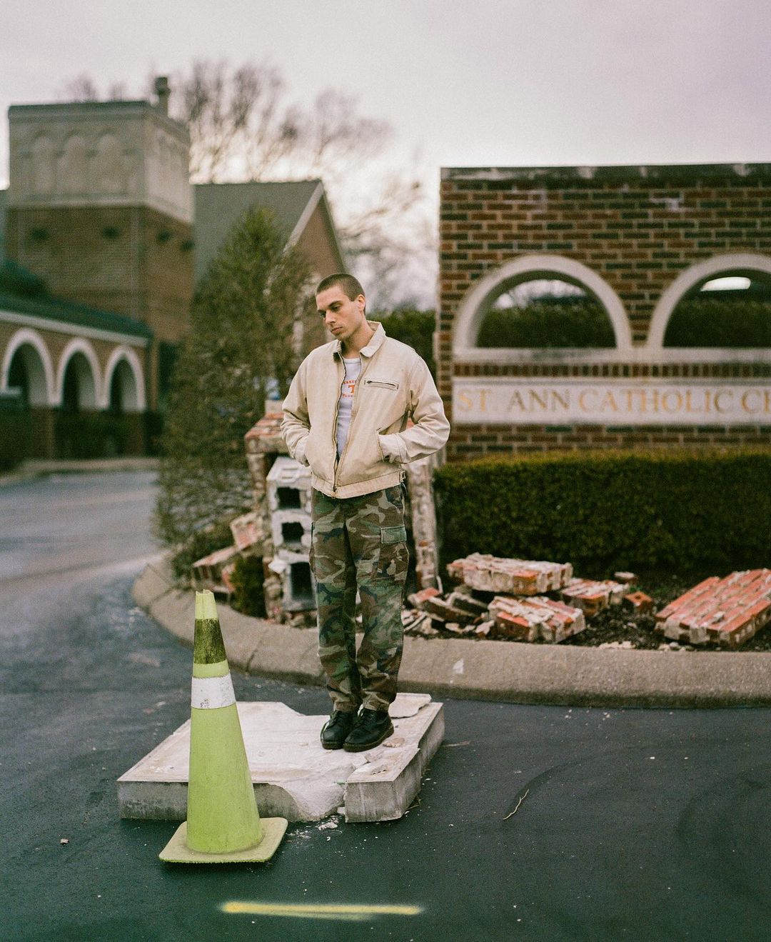 A young adult male stands on a concrete block near a traffic cone, wearing a beige jacket and camouflage pants in front of a church building.