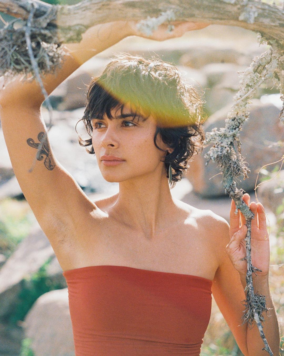 A tanned woman with short dark hair wearing a rust-colored tube top stands outdoors among rocks and a leafy branch in natural light.