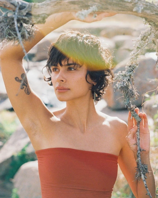 A tanned woman with short dark hair wearing a rust-colored tube top stands outdoors among rocks and a leafy branch in natural light.
