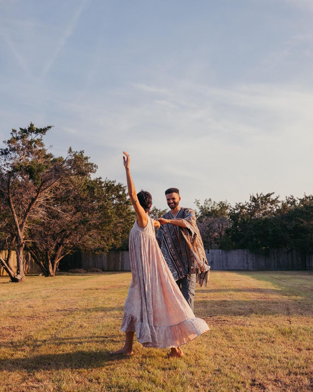 Outdoor shot of a woman in a flowing dress dancing while a man smiles in a yard under a blue sky, showcasing a relaxed, joyful moment.