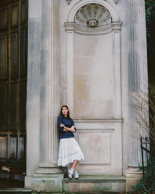 A young woman leans against a stone wall with columns outside, wearing a blue sweater and white lace skirt in natural daylight.