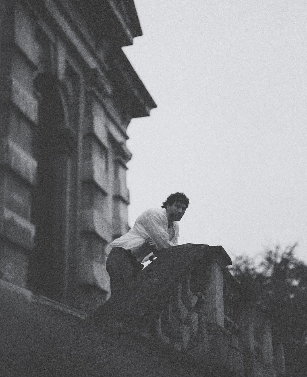 Monochromatic portrait of a man in a white shirt leaning on a stone balcony with trees and an old building in the background.